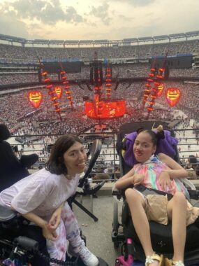 Two women, both seated in power wheelchairs, smile for a photo from their seats at MetLife Stadium. In the background, a large stage is lit up with crowds of people surrounding it.