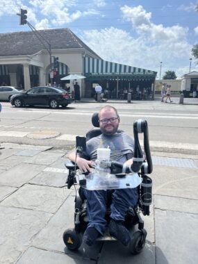 A 30-year-old man smiles for a photo on a stone sidewalk. He's seated in his power wheelchair and appears to be holding a cup in front of him. Behind him is a street and a building with a white and green striped awning. It's a sunny day, and several people are out walking in the background.