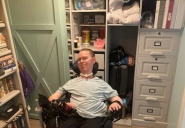 A man in a pale green shirt and black pants sits in a wheelchair in front of a cupboard and file cabinet. At the side are a bookshelf and a light green barn door.