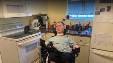 A man wearing a pale green shirt and black pants sits in a wheelchair. He's in a small kitchen, with an oven, sink, and refrigerator close together.