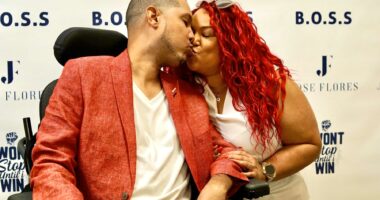 A couple, one a man wearing a white shirt and red jacket, and a woman with red hair wearing a white shirt share a kiss.