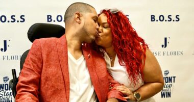 A couple, one a man wearing a white shirt and red jacket, and a woman with red hair wearing a white shirt share a kiss.