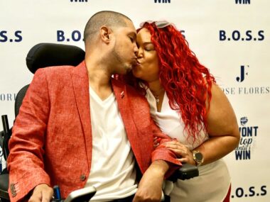 A couple, one a man wearing a white shirt and red jacket, and a woman with red hair wearing a white shirt share a kiss.
