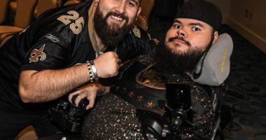 A man poses next to another man, in a power wheelchair, who is wearing a hat.