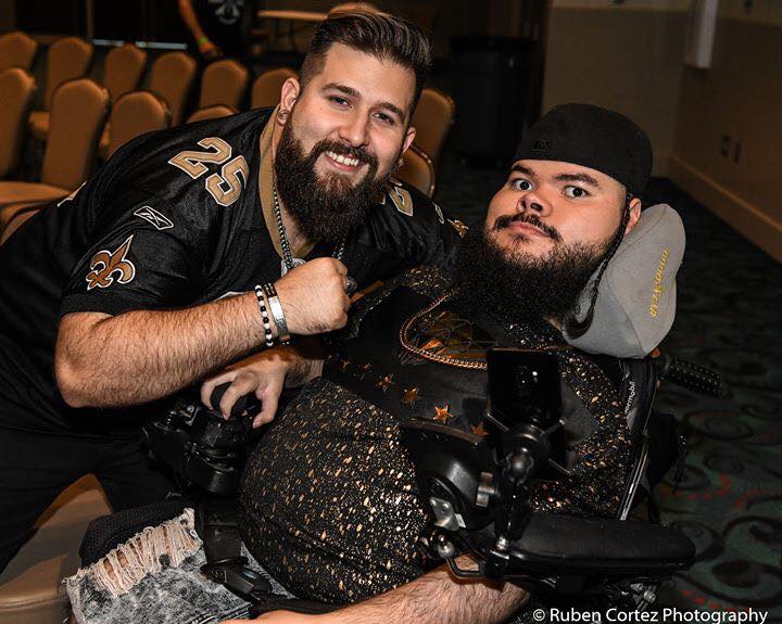 A man poses next to another man, in a power wheelchair, who is wearing a hat.