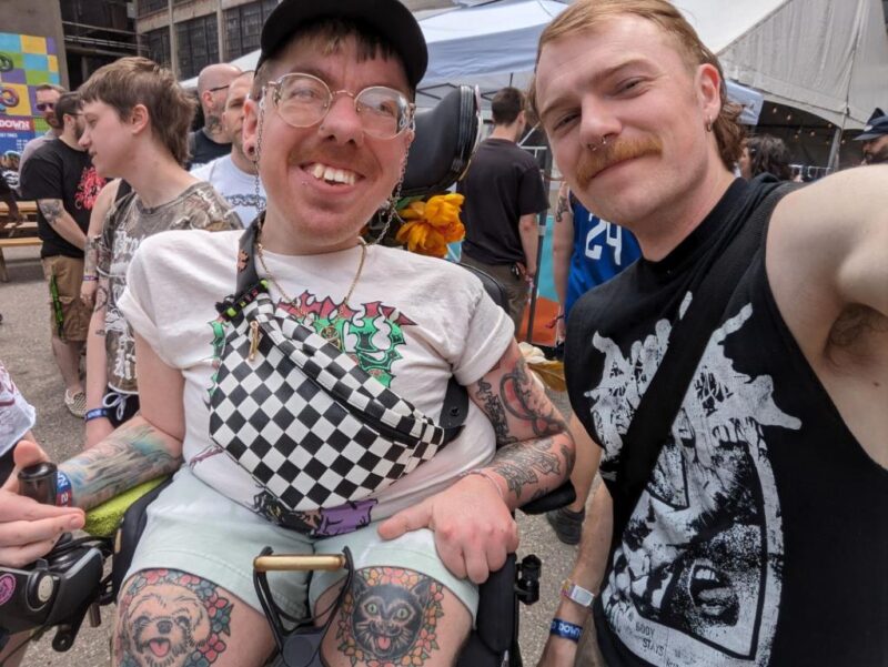 A man with glasses and sitting in a wheelchair poses, smiliing, with another man.