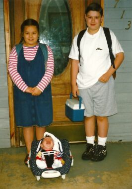 A young girl and boy stand side by side in front of the door to a home in a 1997 photo. In front of them, a baby sits in a carrier wailing and sobbing. 