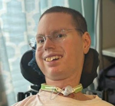 A close-up headshot of a man in a power wheelchair. His head is tilted slightly to the right, he wears glasses, and he is smiling broadly. His tracheostomy is visible toward the bottom of the frame.