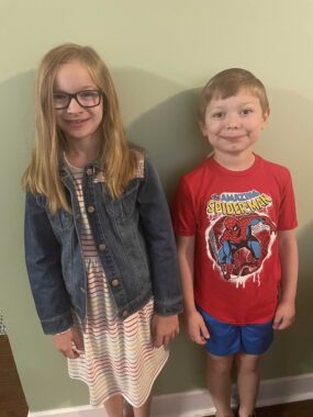 A young girl and boy - siblings - stand next to each other against a wall in a house. The girl, wearing glasses, is smiling coyly, while the boy, in a Spider Man T-shirt stands upright and rigid. 