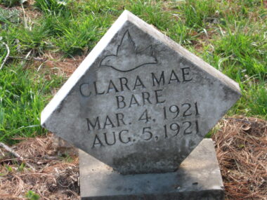 A square headstone at a diagonal angle reads "Clara Mae Bare Mar. 4, 1921 Aug. 5, 1921." It seems brown straw or grass is in a circle around it, with some patchy green grass beyond.