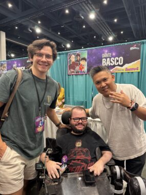 Three men pose for a photo in front of a booth at a convention. The man in the center is seated in his power wheelchair, and his friend and caregiver is standing to his right. The man standing to his left is holding a hand near his face, and all three are smiling. A poster in the background reads "Dante Basco."