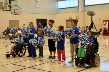 Seven young girls form a row in the middle of a gymnasium. Two of the girls are in wheelchairs, and the other five stand between them. All are holding blue and white pompoms, and most are wearing blue shirts.