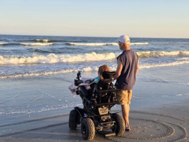 With their backs to the camera, we see a woman sitting in an all-terrain wheelchair and a man standing in a sleeveless blue T-shirt and brown shorts, both facing the ocean waves from shore. Tracks from larger-than-normal wheelchair tires are behind them.