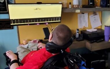 A man sits in a power wheelchair with his back to the camera. He is looking at a long computer screen attached to the wall. Reminder notes or schedules are pinned to the wall to the right. 