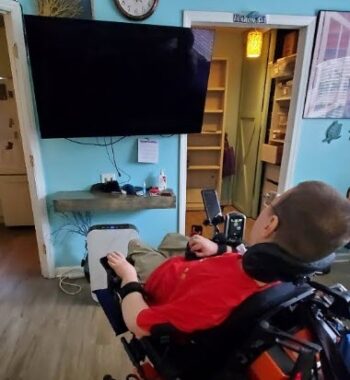 A man in a power wheelchair has his back to the camera while looking at a large TV hanging on the wall. Underneath it is a small shelf. Above it is a large clock. The walls are painting light blue, which goes well with the light-colored wood on the floors and around the doors.