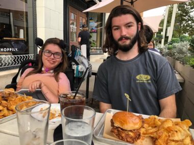 A pair of siblings in their 20s sit on the same side of a table on the patio of a restaurant. The woman on the left is seated in a power wheelchair and wearing a pink shirt and glasses. Her brother is wearing a gray T-shirt and leaning slightly toward her. Burgers, fries, and drinks are on the table in front of them.