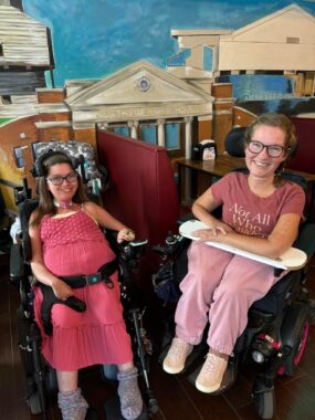 Two women, both seated in power wheelchairs, smile for a photo inside of an ice cream shop. The woman on the left is in a pink sundress, while the woman on the right is wearing a pink shirt and joggers. There are several booths behind them, and there's a mural of a city painted on the wall in the background.