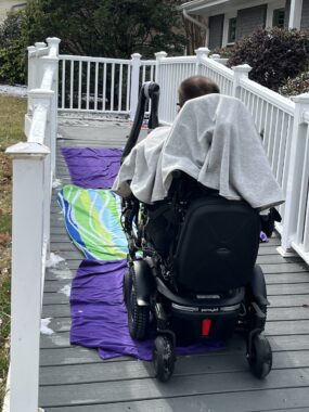 A man in a power wheelchair makes his way up a ramp outside of his house. The center of the ramp is lined with beach towels.