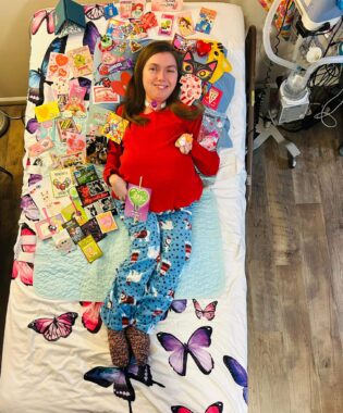 A woman in a red sweater, blue pajama pants, and socks lies on her back in bed, surrounded by a few dozen Valentine's Day cards. The photo is taken from above her, and almost her entire bed is visible. Underneath the cards we can see a white comforter with colorful butterflies, and there is some medical equipment available in the background.