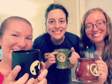 Three woman hold up mugs and lean in for a photo. The mugs are different colors – blue, green, and red – but they all bear the same logo. The women look to be inside someone's house, in a brightly lit room.