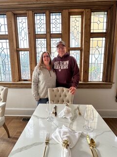 A man in a maroon hoodie and baseball cap stands next to a woman, with his arm around her, as both are posing in front of a set of antique windows. The luxurious table set in front of them indicates to the viewer that they're at a fancy restaurant.