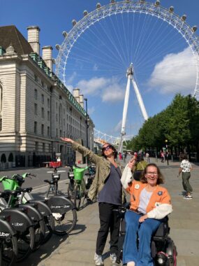 Two women pose for a photo on a sidewalk in front of the London Eye. One woman is seated in her power wheelchair, her right arm extended as if pointing toward the Ferris wheel. The other woman is standing next to her, with both arms outstretched and face angled toward the sun, like she's soaking it all in. It looks like a beautiful sunny day, and both women are smiling broadly.