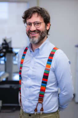 A professional photo shows a man, from his waist up, standing with his hands clasped behind his back. He's wearing a long-sleeve, blue, button-down shirt under colorful, striped suspenders, as well as round glasses. He has brown hair and a short, graying beard, and is smiling happily at the camera.
