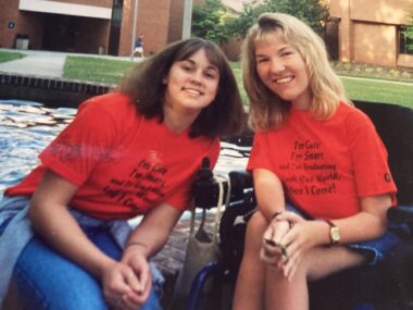 Two young women wearing matching red T-shirts smile and lean in for a photo. The woman on the right is siting in a power wheelchair while the woman on the left is sitting on the edge of a fountain. It looks like they're on some sort of campus, perhaps their high school, as there are large brick buildings in the background overlooking a small lawn. 