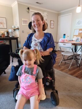 A woman in a wheelchair sits behind a grinning young girl in blond pigtails, pink overalls with short pants, and a gray T-shirt. The woman, with short blondish-brown hair, is wearing a white floral-print shirt with a blue shirt over it. They're indoors in what appears to be a living room, with a simple table and cork board to one side.