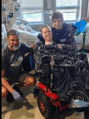 Three people are in a hospital room in front of a bed. The one at center is in a power wheelchair; he's flanked by a bearded man, squatting at left, and an older woman over his shoulder at right.