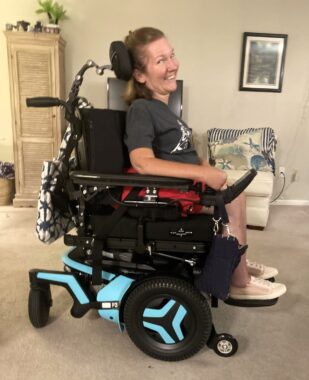 A woman with dark blond hair sits in a much larger wheelchair. She's in a home with carpet.