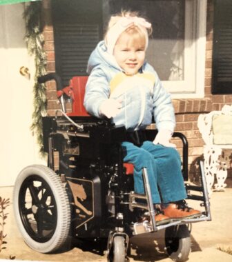 A young blond girl sits in what appears to be a small power wheelchair. She's outdoors in front of a brick house with a white door.