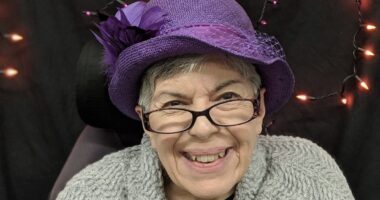 A headshot shows an older woman smiling and wearing a purple hat and glasses.