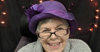 A headshot shows an older woman smiling and wearing a purple hat and glasses.