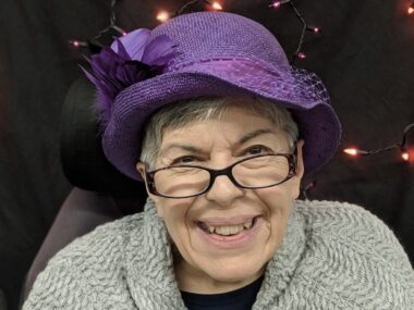 A headshot shows an older woman smiling and wearing a purple hat and glasses.