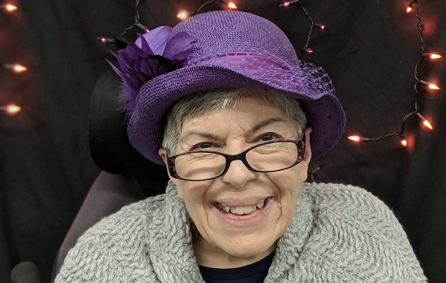 A headshot shows an older woman smiling and wearing a purple hat and glasses.