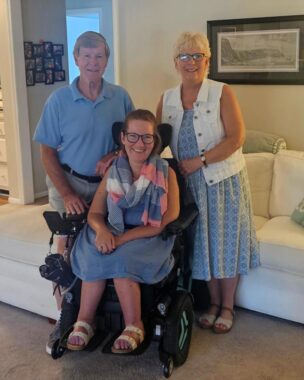 In a living room in front of a white sofa, a woman with reddish-brown hair wears a blue skirt with a blue, red, and white scarf and sits in a wheelchair. Behind her are an older man and woman. He wears a blue polo shirt and has reddish-brown hair; she wears glasses, a blue dress, and a white vest and has blond hair.