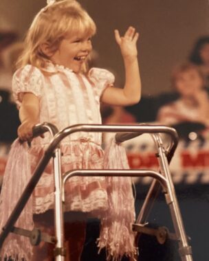 A young girl wearing a pink dress smiles and waves to an audience. She has blond hair and is leaning on a small walker for support.