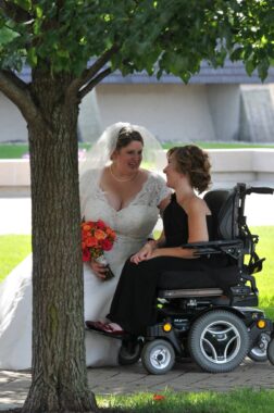 A woman in a black dress sits in a power wheelchair on a sidewalk outside. Another woman wearing a wedding dress and veil, and holding a bouquet of orange flowers, sits beside her. They are leaning toward each other and smiling, as if in conversation.