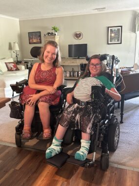 Two young women smile brightly in a home setting. Both are in power wheelchairs and have their chairs angled slightly toward each other, indicating a closeness between them.