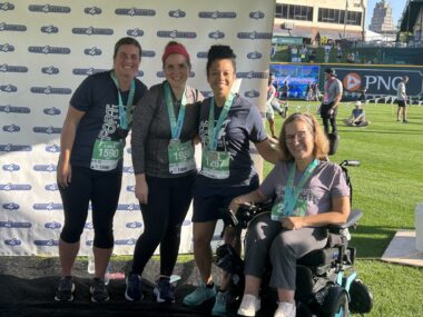 Four women — three standing, one in a wheelchair — are shown in racing outfits with medals draped around their necks.