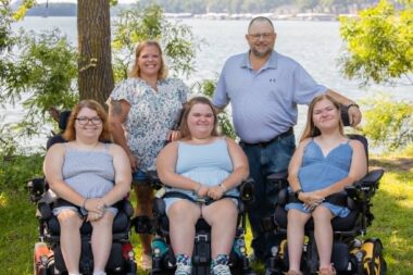 A family poses for a family photo in front of a lake or other similar body of water. They are in the shade of a tree. Mom and Dad stand in back, and three girls are in wheelchairs in the front fow. They're all wearing light colors, mostly blue, to keep cool on what is probably a warm day. 