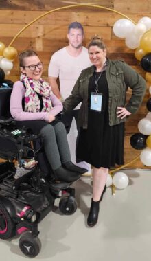 Two women are beaming with smiles as they pose in front of a Tim Tebow cutout, with white, gold, and black balloons. Both are casually dressed, and the woman on the left is in a power wheelchair while the woman on the right stands with one arm on the chair and the other on her hip. 