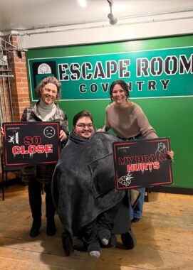 Three people post for a photo in front of an escape room sign. The man in the center is seated in a wheelchair and covered with a large gray hoodie, while two women — his mom and his best friend — stand on either side of him. His mom holds a sign that says "So close" while his friend's sign reads "My brain hurts."