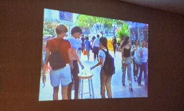 A video reel projected onto a wall shows a bustling pedestrian walkway in an urban setting in the 1990s. Young onlookers gather around a wooden stool almost in the center of the frame. 
