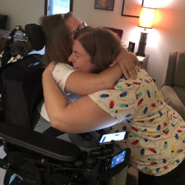 Two friends laugh while sharing a bear hug. One of the women is seated in a power wheelchair, and the other is kneeling in front of her so their shoulders are at the same height.