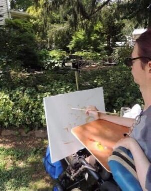 A man in a wheelchair works on a painting outside in the shade. He is parked on the grass and looking at lush green foliage.
