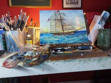 A variety of art supplies sit on a desk inside a studio. Propped up next to a palette and set of brushes is a painting of a ship on the ocean. 