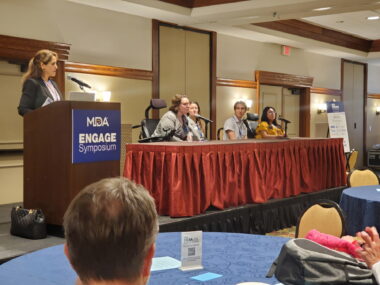 A woman stands at a podium with a plaque reading "MDA Engage Symposium." A table is seen to the right, with a group of people behind it and one person speaking. 