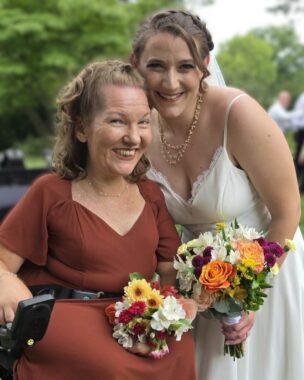 A maid of honor and a bride, both holding bouquets, smile for a photo. The maid of honor is seated in a power wheelchair while the bride stands beside her.